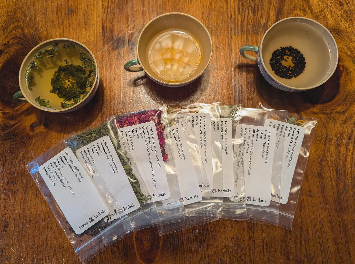 A wooden table displaying three cups of tea and a row of labeled herb packets. The cups show steeping herbs, one with green leaves, another with creamy white liquid, and the last with small dark seeds. The herb packets feature printed labels detailing their names and uses.