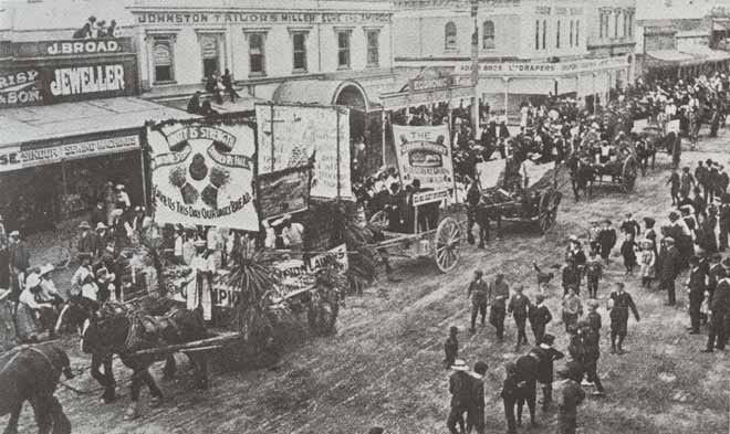 Old black and white photograph of a parade down a main street. There are floats pulled by horses with banners saying things like "Unity is strength". There are people lining both sides of the street, watching the parade.