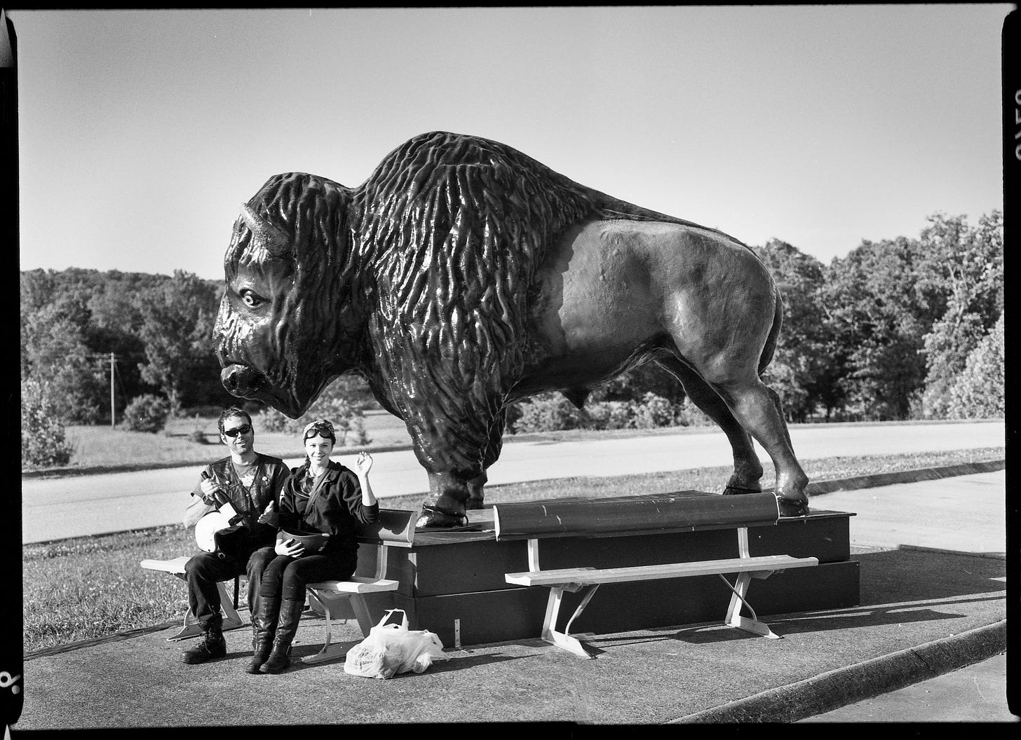 Amber and Derrick sit on a bench in front of a large statue of a buffalo. They make funny faces to the camera. 