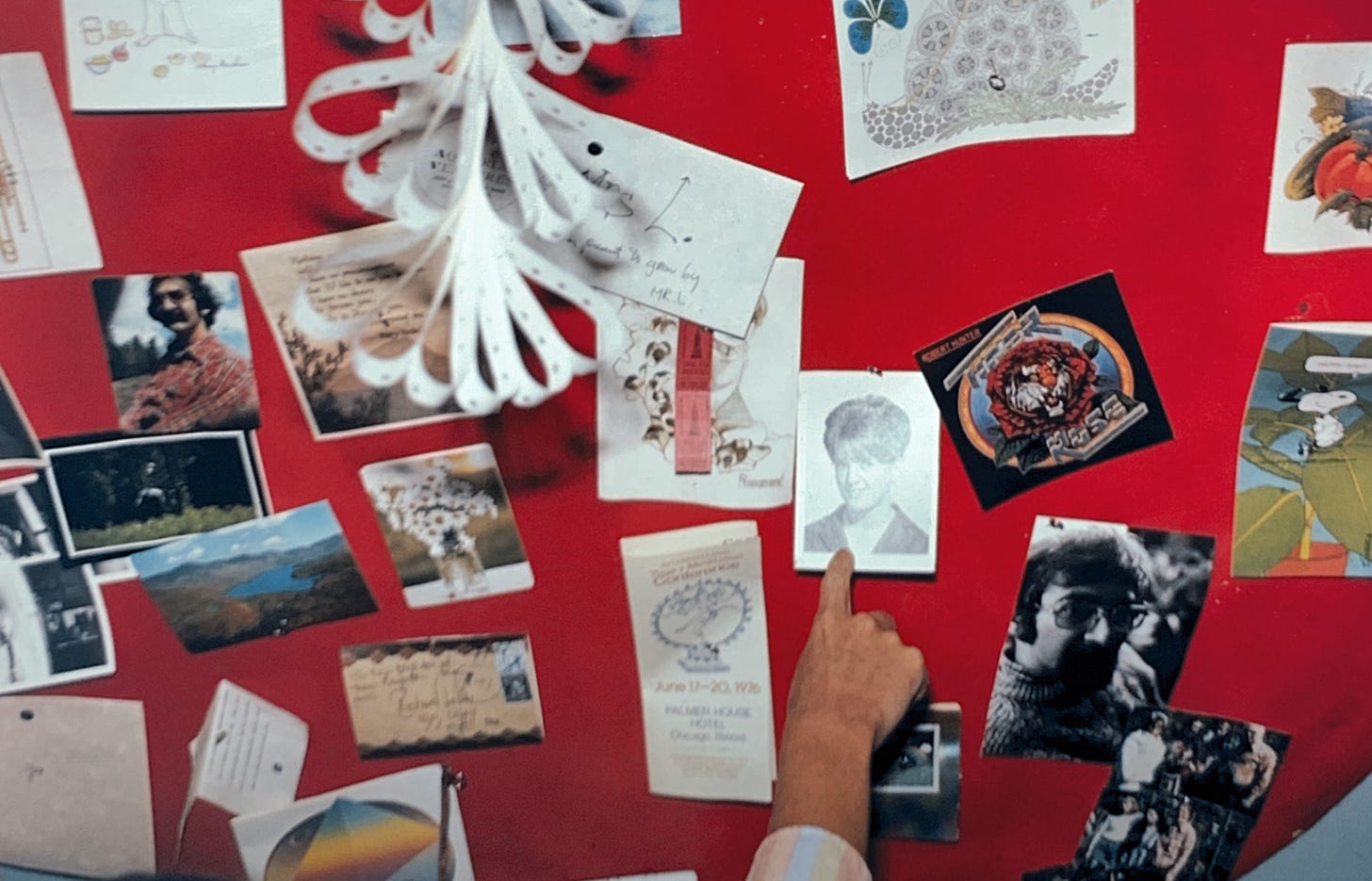 red background with many photographs, postcards, notes with a hand coming from out of frame pointing at a black and white photograph of a senior photo