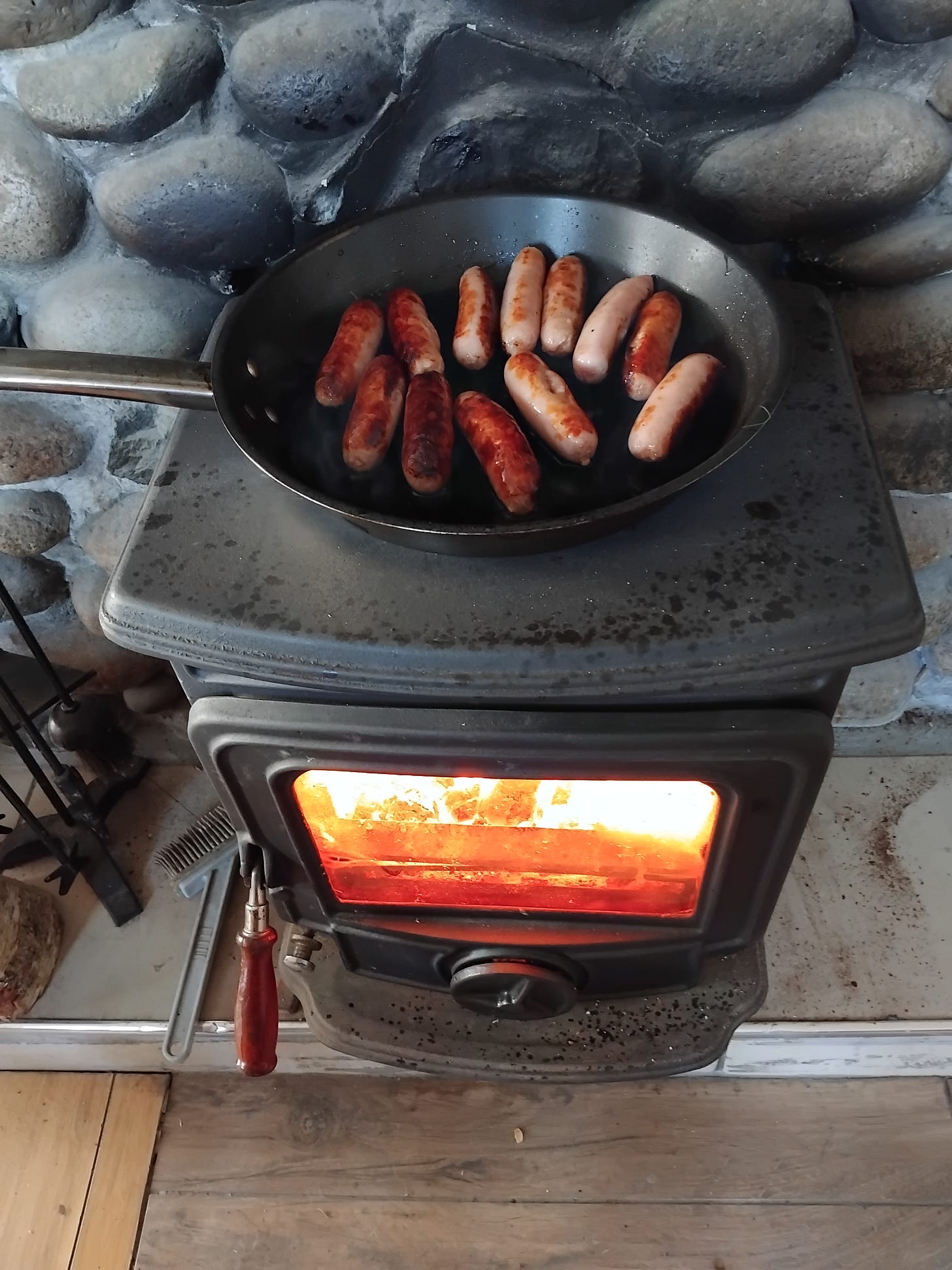 12 sausages are cooking on a pan on top of a wood burning stove. They are crisping perfectly. 