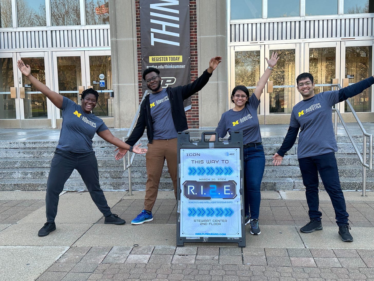 Students in Michigan Robotics t-shirts pose in front of a sign saying "This way to RISE"