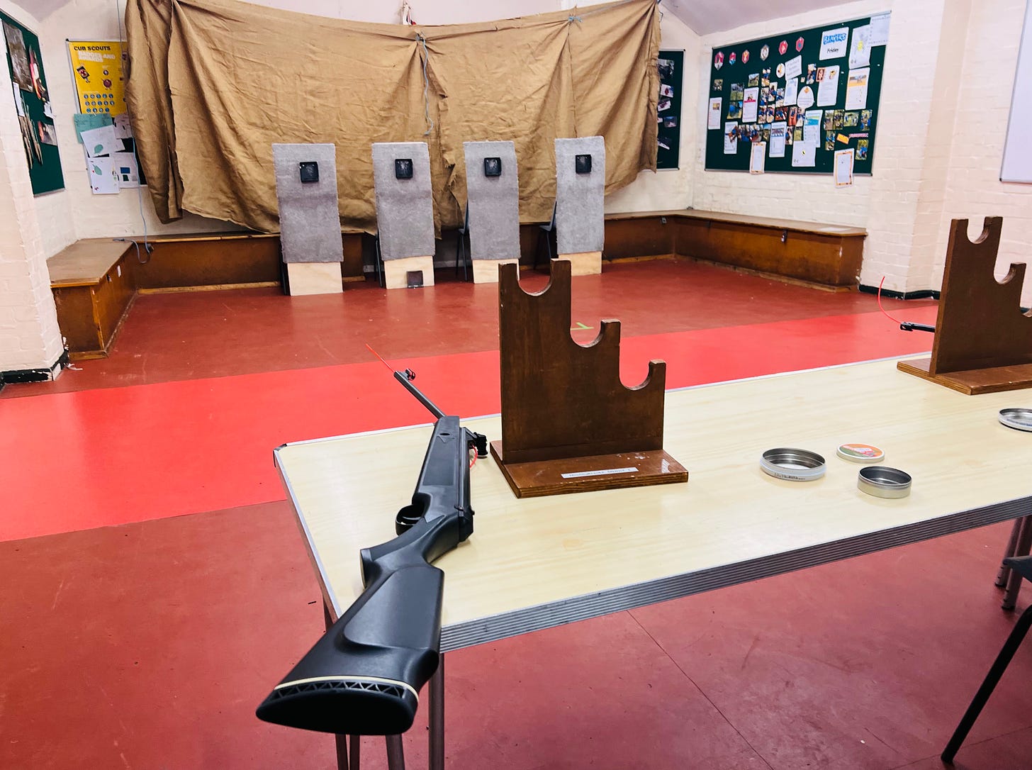 An air rifle sits on a table in a scout hut. four targets can be seen in the distance.