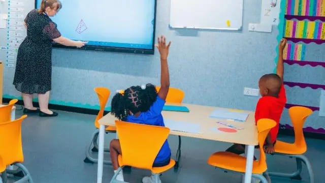Two children, a girl in a blue T-shirt and boy in a red T shirt, sit on orange chairs at a table in a classroom. They hold up their hands to answer a question being asked by the teacher wearing a black patterned dress as she writes on a white board at the front of the classrom.