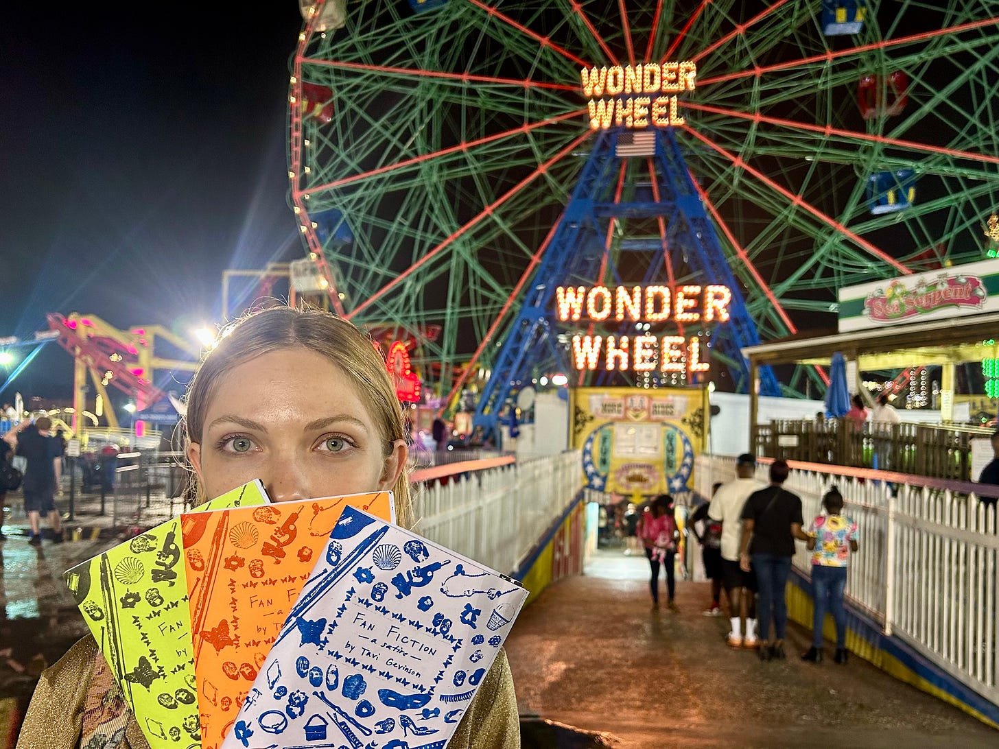 Photo taken at night of Tavi Gevinson in front of a ferris wheel with bright lights that read “Wonder Wheel.” Tavi is in the left corner. Her face is partially obscured by the three copies of the zine “Fan Fiction” that she is holding up. Each copy is printed in a different color: yellow, orange, lilac with blue ink.  