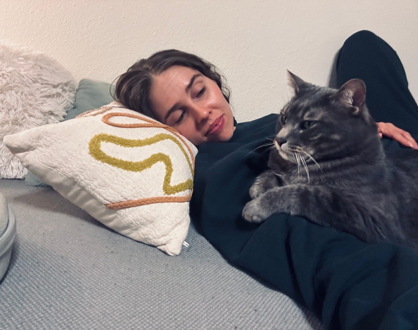 Kaitlyn reclines on a bed with a gray cat on her chest