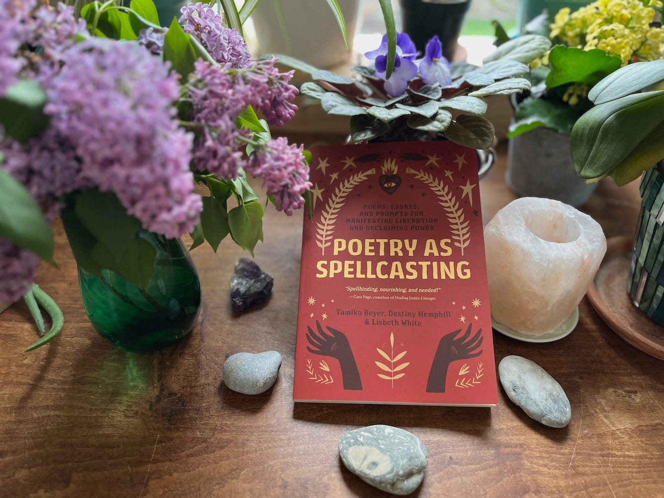 A copy of Poetry as Spellcasting among lush, flowering houseplants, cut lilacs, stones, and crystals.