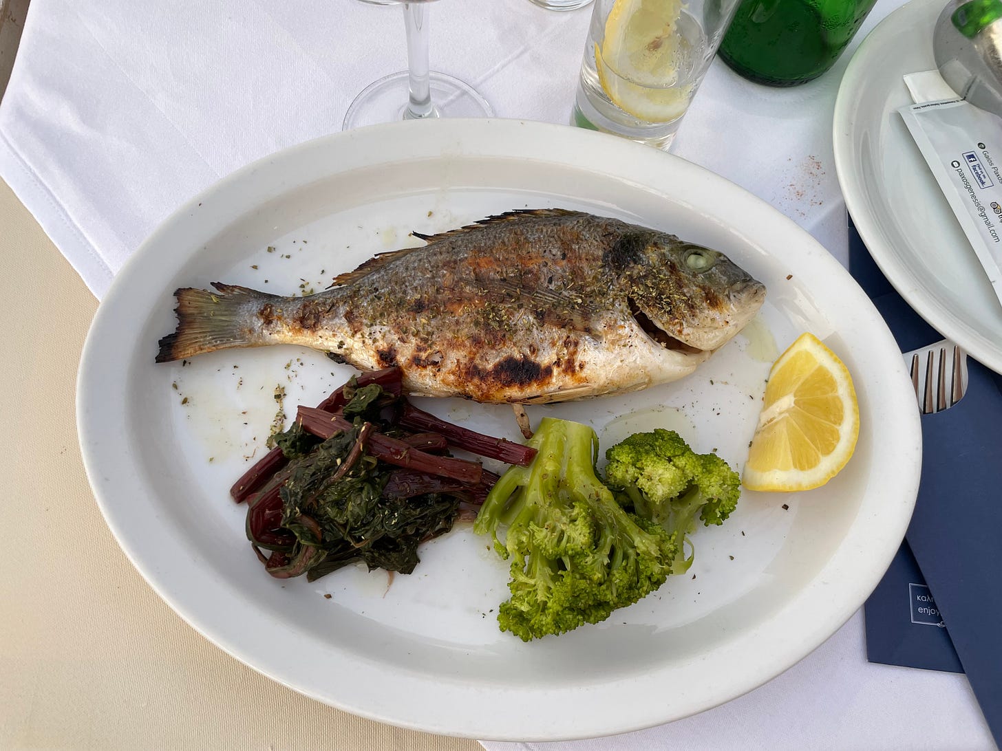 A color photo of a dinner plate with a whole grilled fish, cooked chard, broccoli, lemon wedge. The stem of a wine glass and a glass of water behind on the tablecloth.