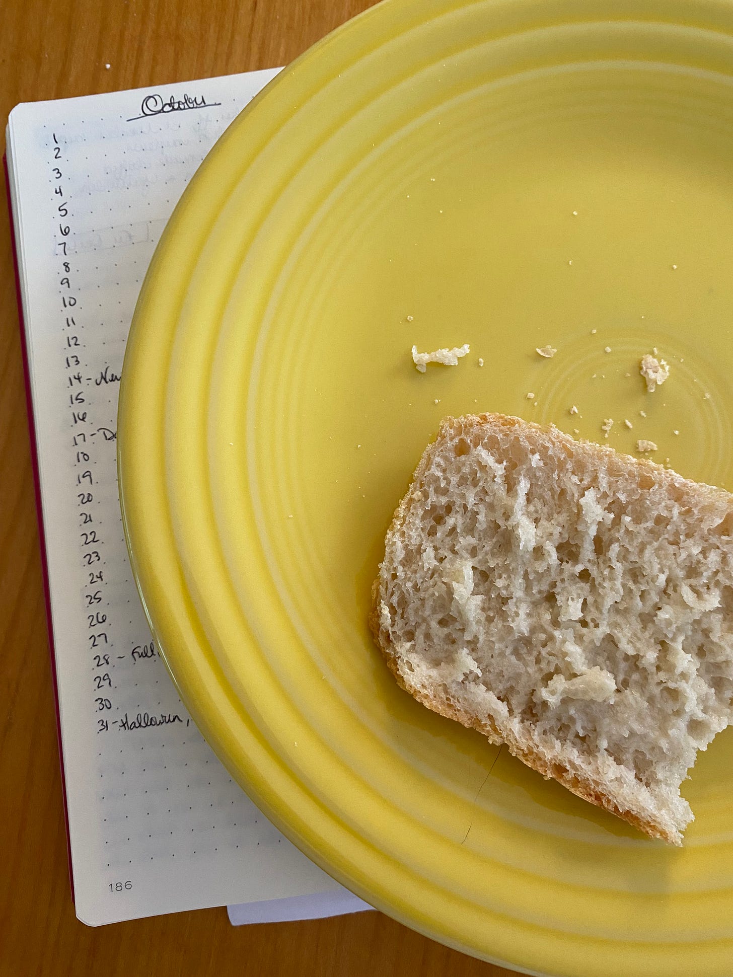 A slice of bread with butter on it has a bite taken out of it. It sits on a yellow fiestaware plate, which sits on top of a notebook open to a page that says "October"