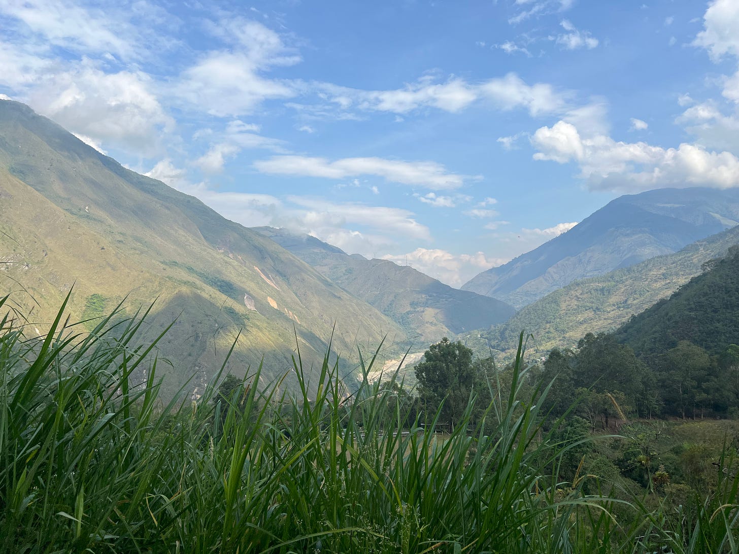 a vista of mountains with a river through them against a partly cloudy sky. there's grass in the lower quarter of the image.