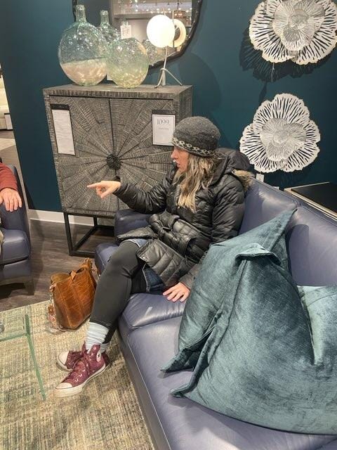 Julie, a white woman, is wearing a winter coat and hat but is sitting inside on a blue leather couch in a furniture store. There are enormous blue velvet couch pillows next to her, and, on the wall, huge metal flower sculptures. She is pointing, because she's in the middle of a conversation. You can see the arm of her husband almost out of the photograph. He is sitting in a leather chair.