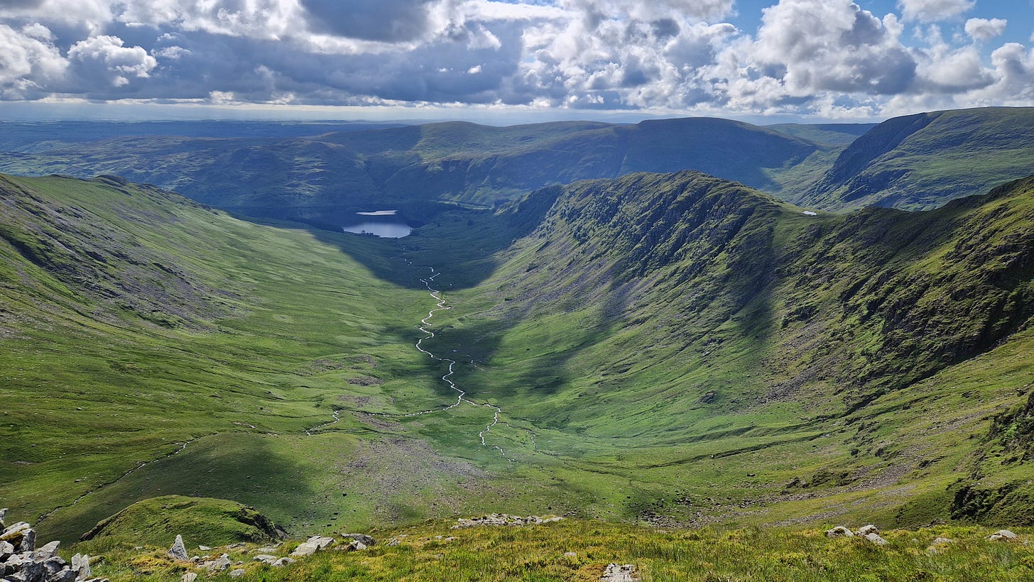 A large valley with stream running towards a reservoir. Steep ridge to right