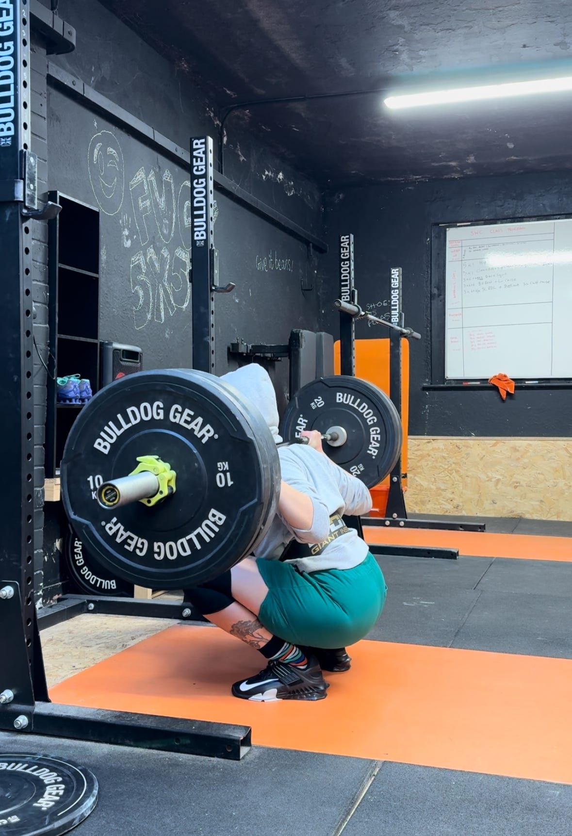 Image shows Pennie squatting 75kg to depth on an orange platform in a black room. They're wearing black Nike lifters, green shorts, funky socks and a grey hoody. They have a leather lifting belt on and the wall in the backhground has "fuck 5x5" written in chalk