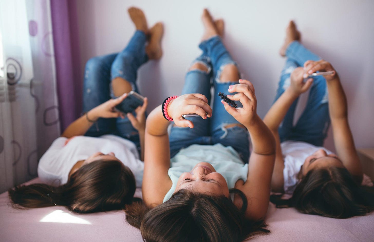 Three young female people lie on their backs, with their feet up the wall. They are all looking at phones.