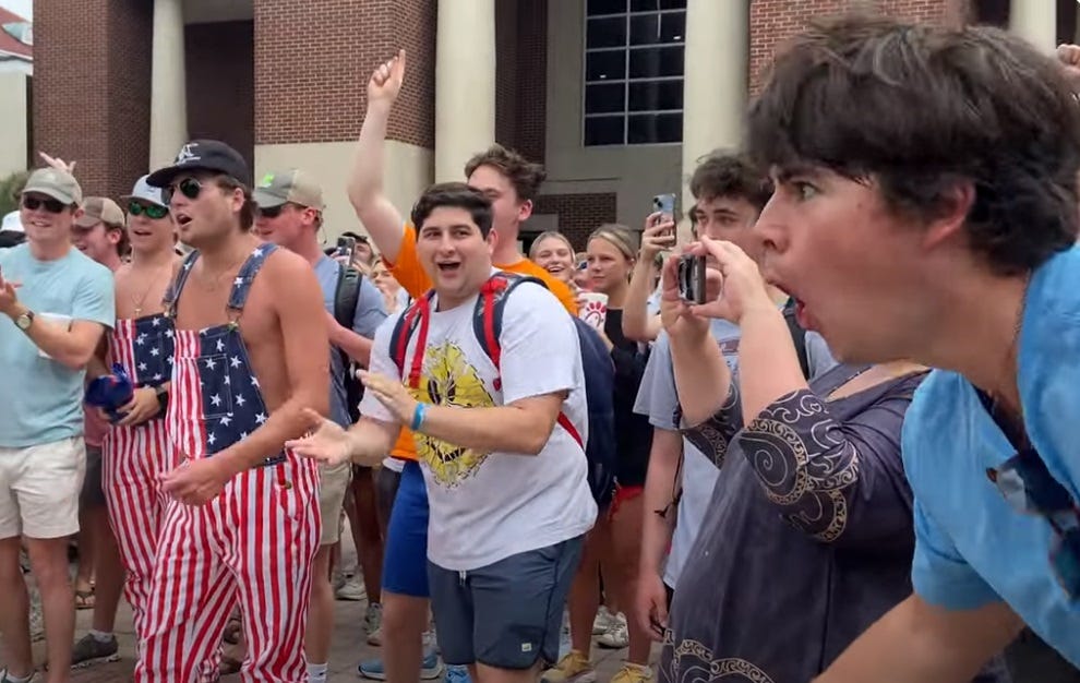 video screenshot of an all-white crowd jeering at a Black woman (offscreen in this frame). Two frat jerks wear US-flag printed overalls with no shirts. at the far right of shot, an asshole in a blue shirt has his lips pursed to make monkey noises.