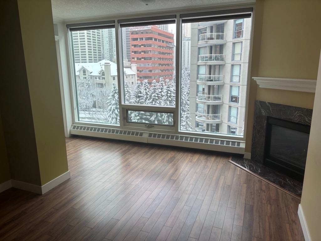 View of an empty living room without any furniture. The floor to ceiling windows show a snow-covered downtown view. The walls are a light olive colour, the hardwood floors dark wood and the fireplace has a black marble-looking hearth.