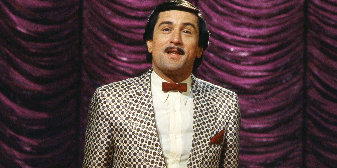 Robert De Niro as Rupert Pumpkin in The King of Comedy. He's standing in front of a purple curtain wearing a garish patterned suit with a bowtie, addressing the audience.
