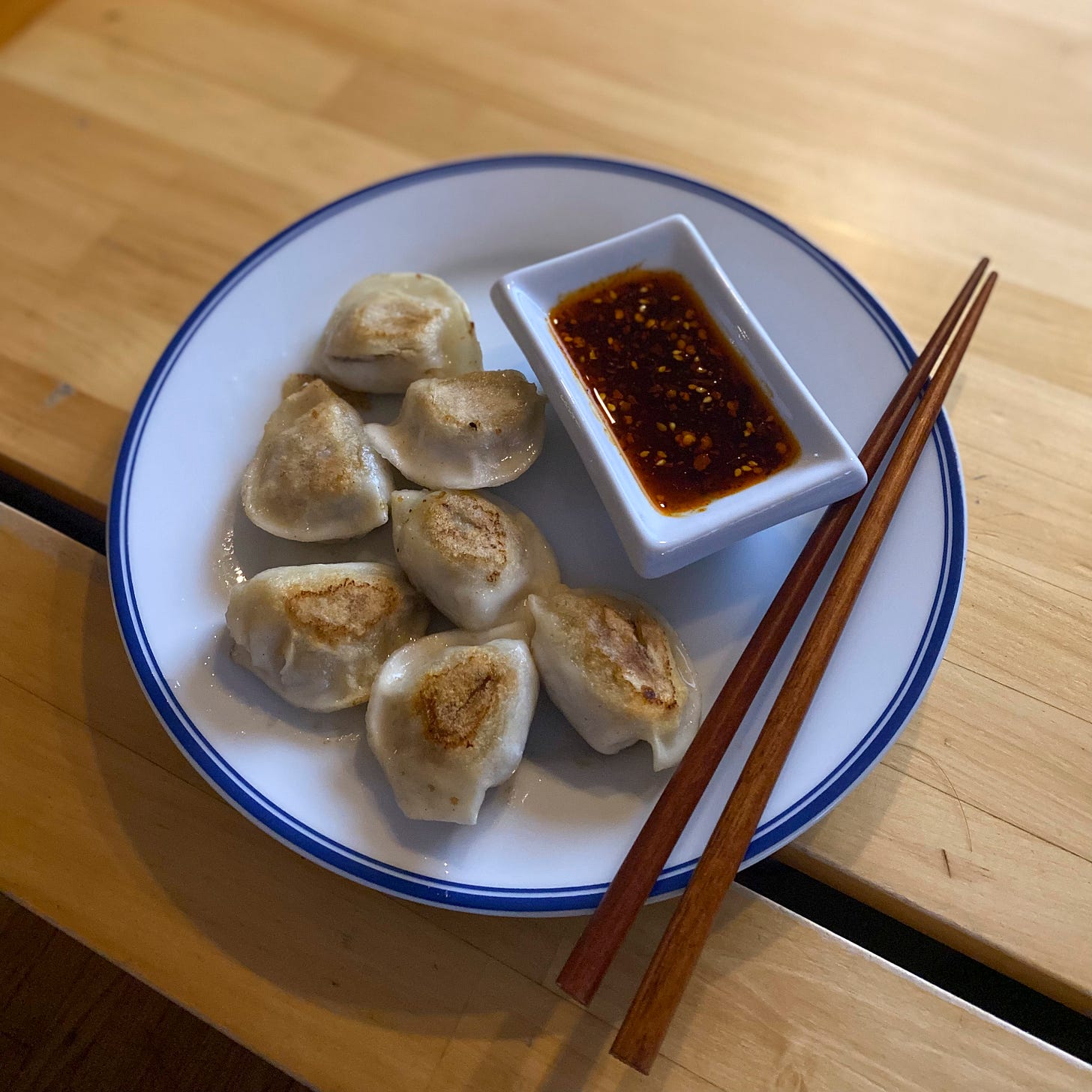 A white plate with a blue rim holding seven half-moon shaped dumplings next to a white rectangular ramekin of dumpling sauce mixed with chili oil. Wooden chopsticks rest at the edge of the plate.