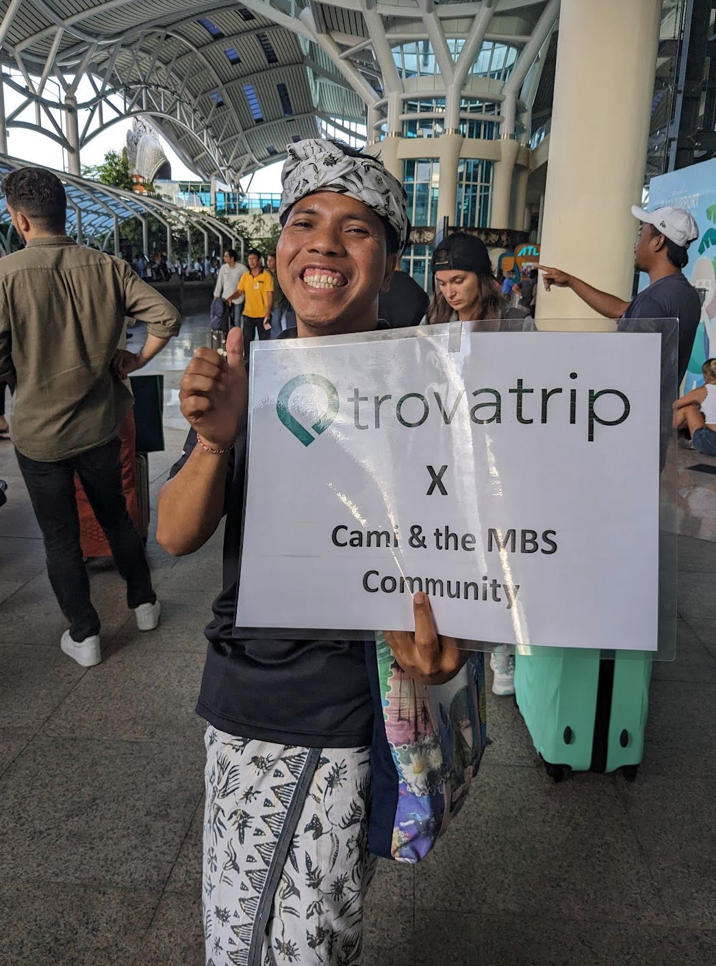 Sana smiles wide enough to show his gums. He wars a white and black headdress, black shirt and white and black sarong. He holds up a sign that reads, "trovatrip x Cami & the MBS Community"