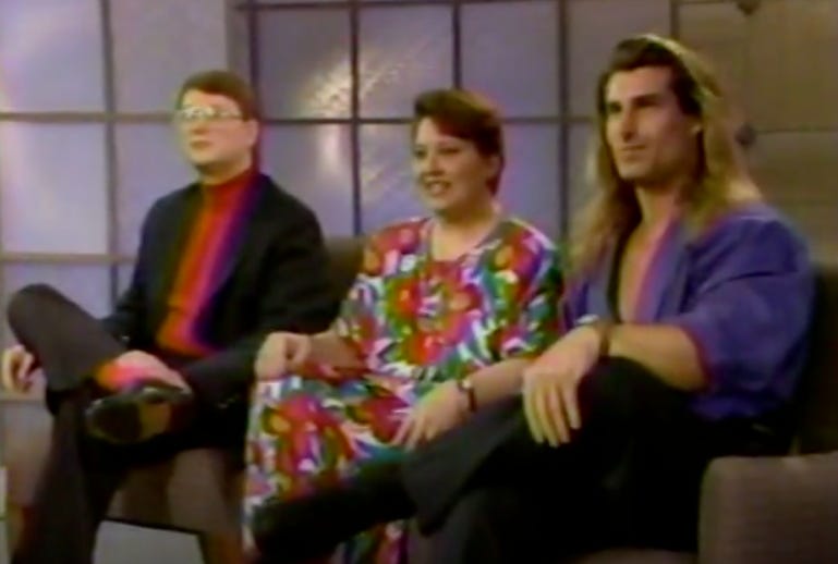 This is a screenshot from Maury in 1992. On the left is Scott Wulf, he has large glasses and is wearing a suit and red turtleneck. In the middle is his wife Ellen, who is wearing a long floral dress. On the left is Fabio, who is wearing black slacks and a blue button-up shirt. The buttons on his shirt are only half-buttoned. 