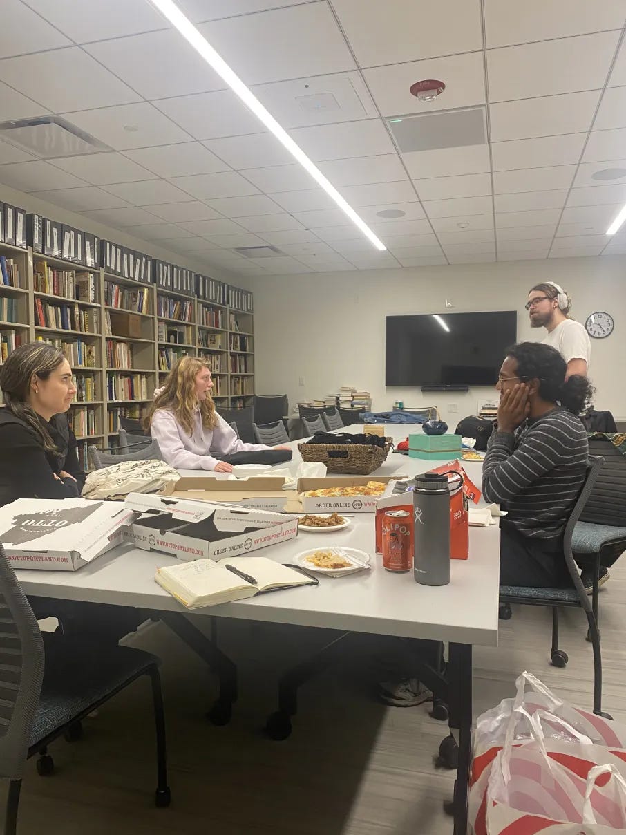 Four students are surrounding a long table, mid conversation.  Pizza boxes, food, and beverages are visible on the table.  The group is located in the Cookbook Library.
