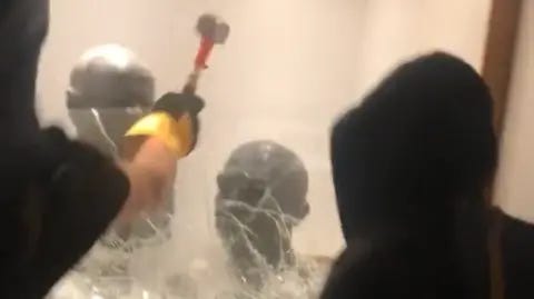 Palestine Action screengrab of two dark-hooded people using a hammer to break a display case with two busts of Chaim Weizmann