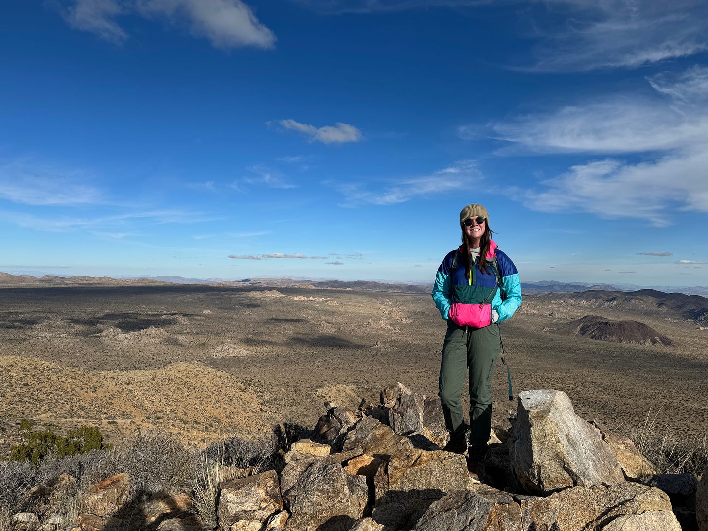 A woman with brown hair and light skin stands among rocks, which are above a vast desert below. She is wearing sunglasses, a brown beanie, green pants and a multi-colored windbreaker. The sky is blue with few clouds.