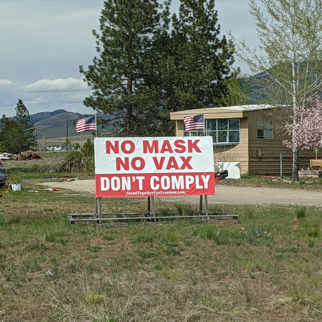 A banner reads NO MASK, NO VAX, DON'T COMPLY, flanked by two waving American flags, in front of a mobile home trailer in a rural setting
