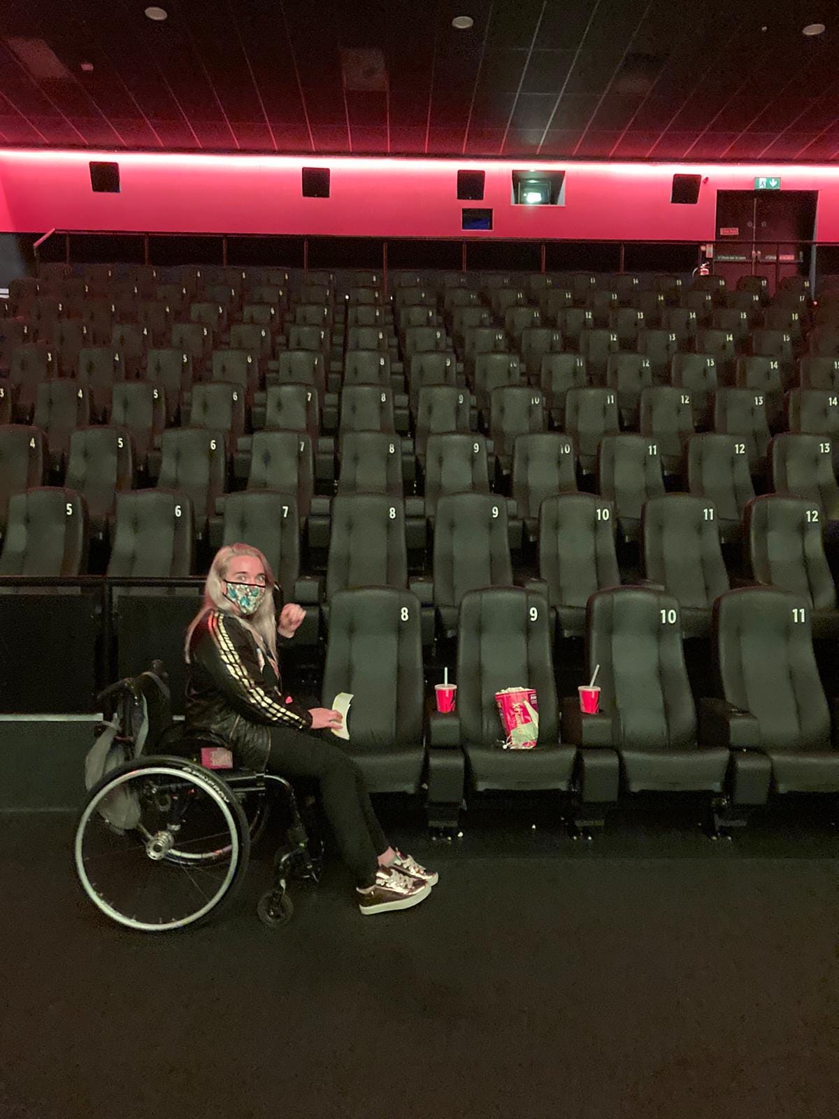 Louise sits in her wheelchair in front of an empty cinema screen, with her coke and popcorn placed on the seats. 