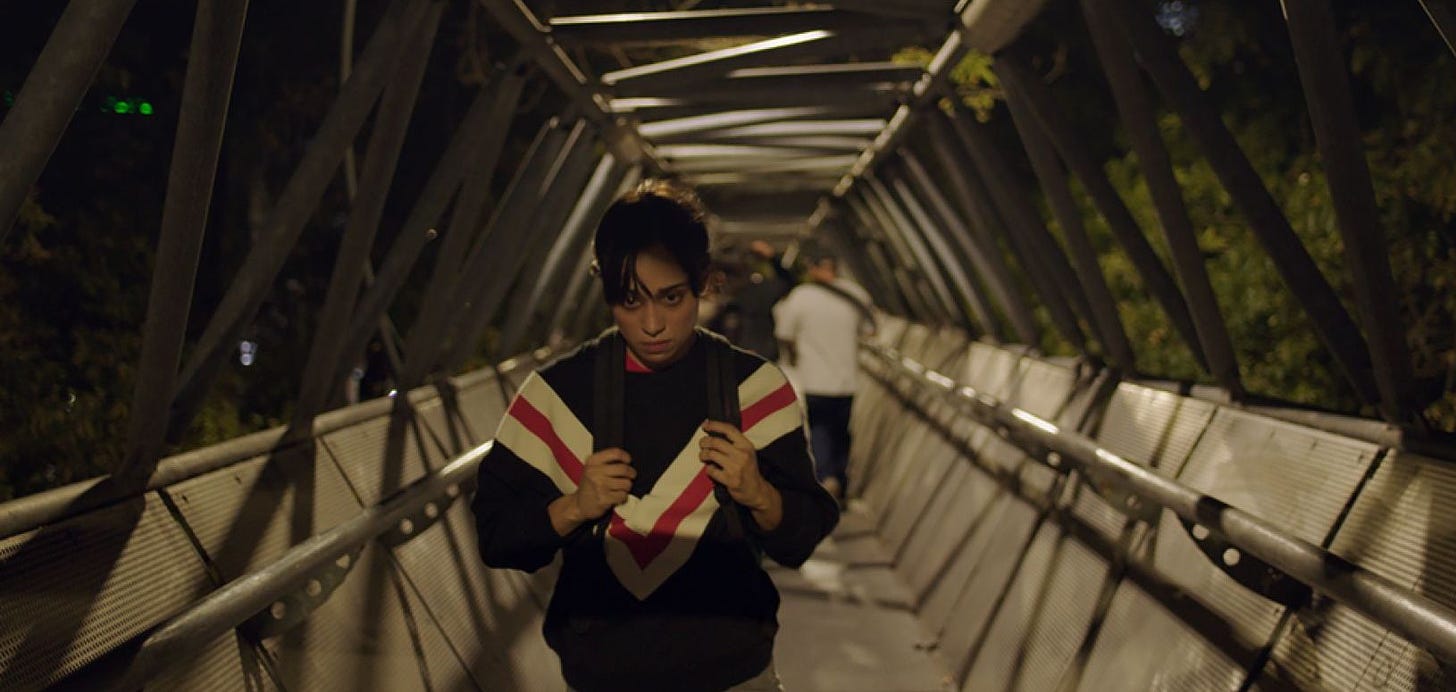 A young woman with dark brown hair pulled back, is walking towards the viewer on a cage-like overpass. She looks sad and serious as her hands clutch her backpack straps against her torso, clad in a black sweatshirt with a thick white V with a red stripe through it. 