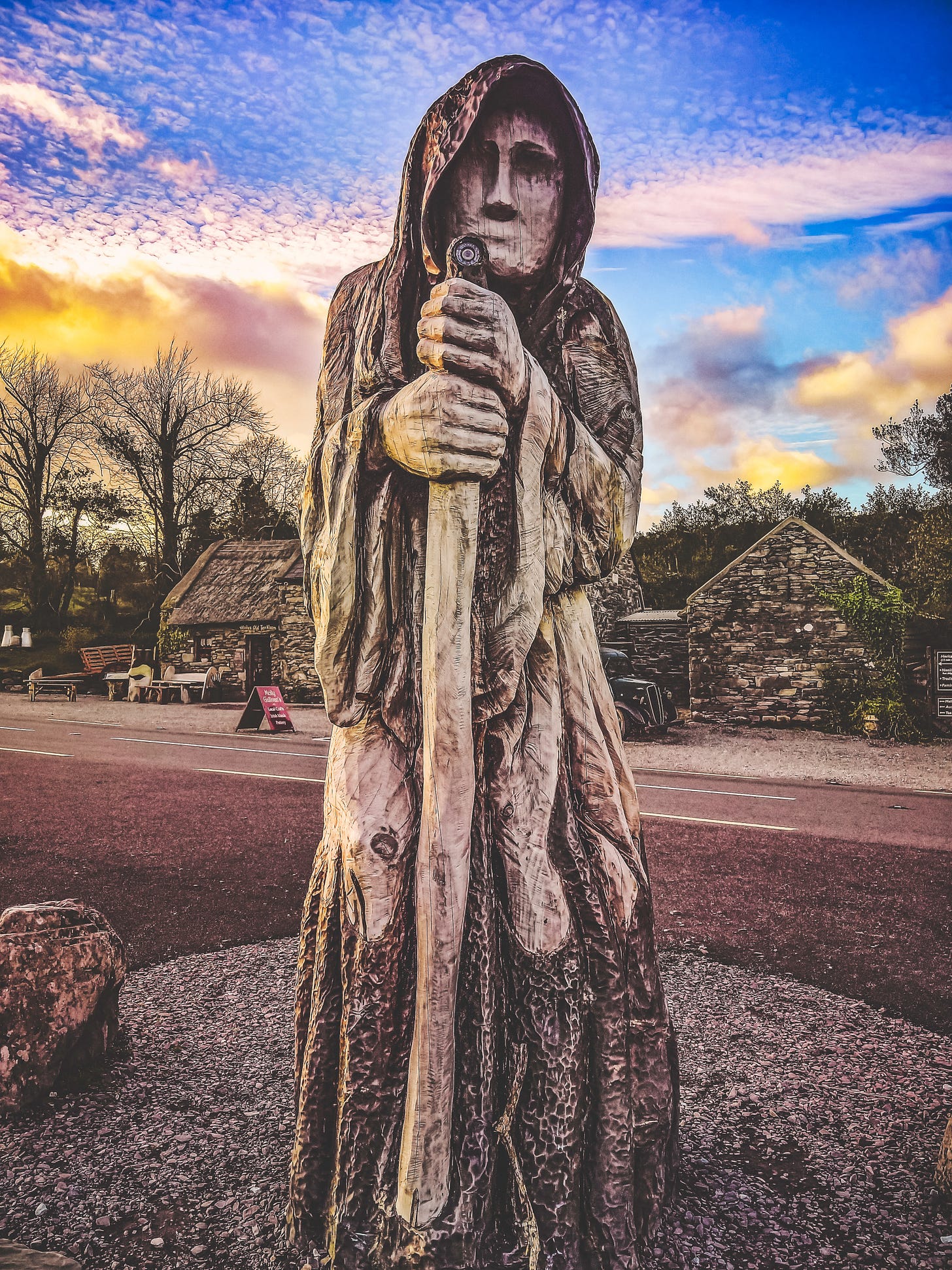 Statue of a crone against a blue and yellow clouded sky in a village with stone houses along a paved road.
