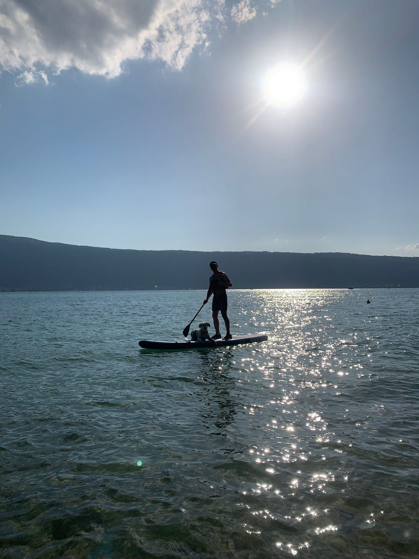 A man paddleboarding with his dog on Lake Annecy, France, under a bright sun and clear sky, with sparkling water and distant hills in the background.