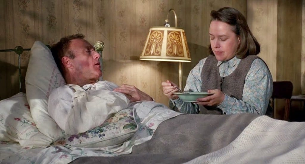 A still from 'Misery'. Paul Sheldon (James Caan) is in bed, wrapped in dated floral bedsheets, being tended to by Annie Wilkes (Kathy Bates). She sits on a wooden chair by the bedside, a lamp lighting the scene next to them. She is feeding him something from a bowl. He wears a nightshirt and she wears a grey long sleeved shirt and dark grey pinafore, with short brown hair. 