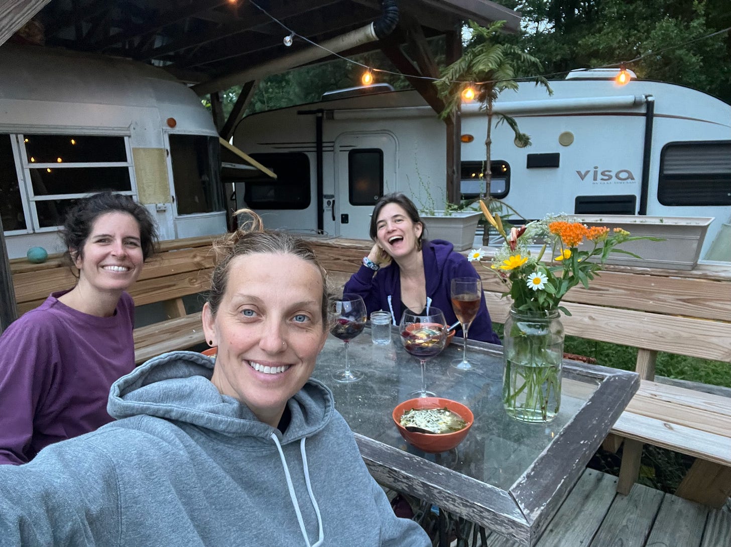 Three brown haired people in sweatshirts smiling and laughing while sitting around a table with wine and soup and fresh flowers, on a wooden deck, in front of two camper vans with twinkle lights strung above.