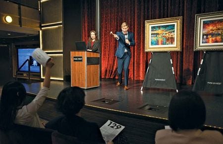 a man standing at the front of a room with a gavel pointing to an audience member with a piece of art behind him