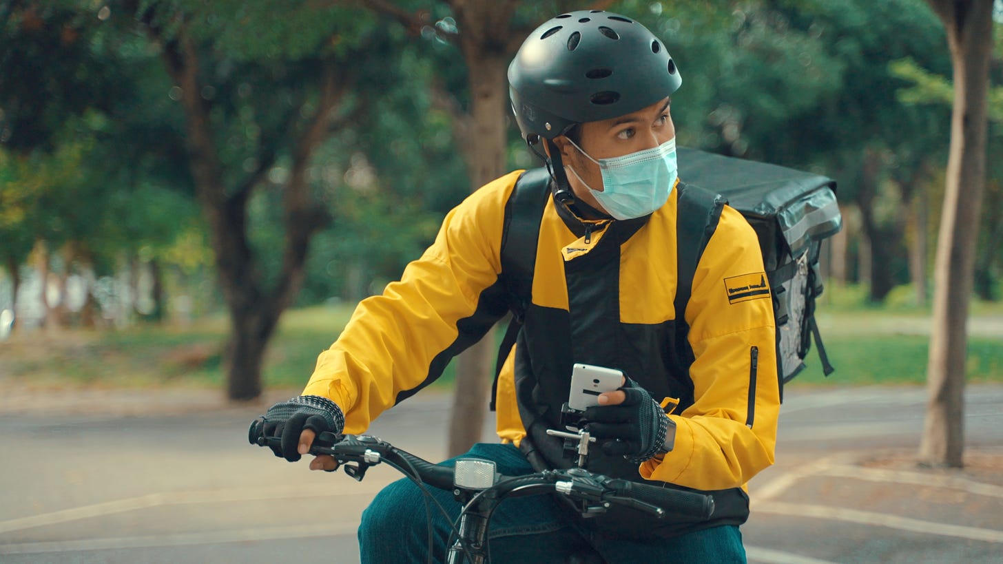 a delivery man on a bicycle wearing a facemask and holding a smartphone navigates a park