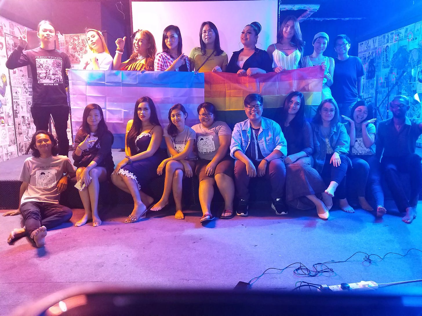 Group photo on a stage with pink and blue lighting. Folks on the back row are holding the trans and rainbow flags.