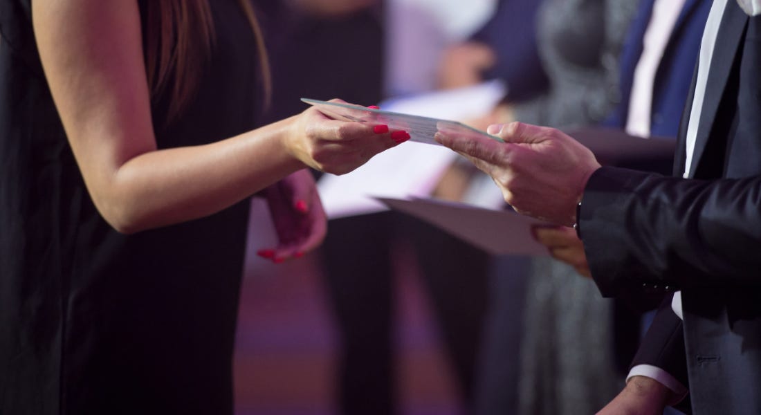 A person wearing a black dress receiving a white letter from someone in a black suit A person wearing a black dress receiving a white letter from someone in a black suit