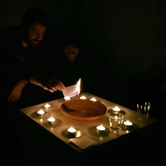 Two men sit in the darkness, lit by ten tea candles around a wide clay bowl. One man is holding a flaming index card above the bowl.