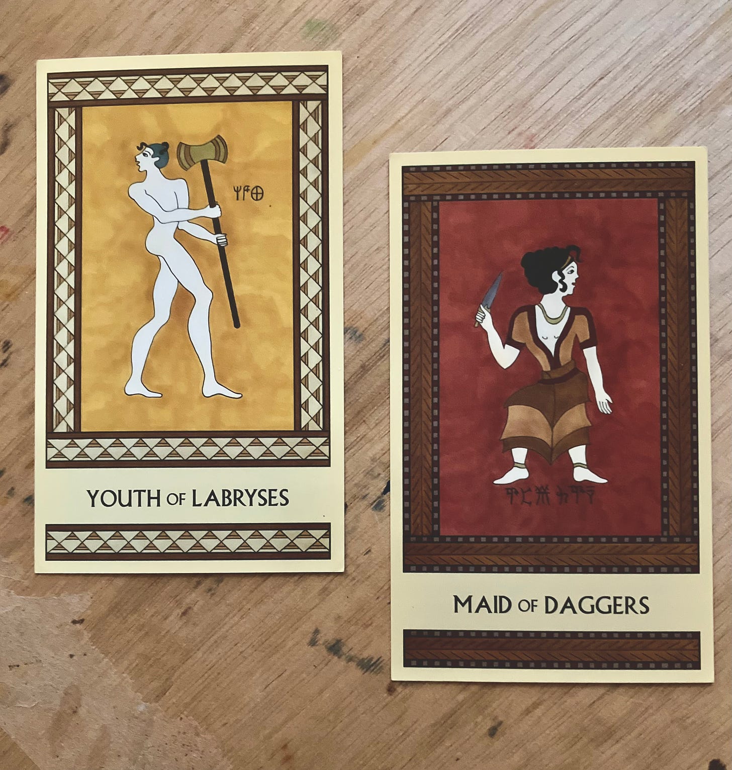 Two Minoan Tarot cards on the stained wooden surface of an art desk. The Youth of Labryses is in shades of gold and cream. It shows a naked young man holding a large labrys. The Maid of Daggers is in shades of dark red and brown. It shows a Minoan girl holding a dagger.
