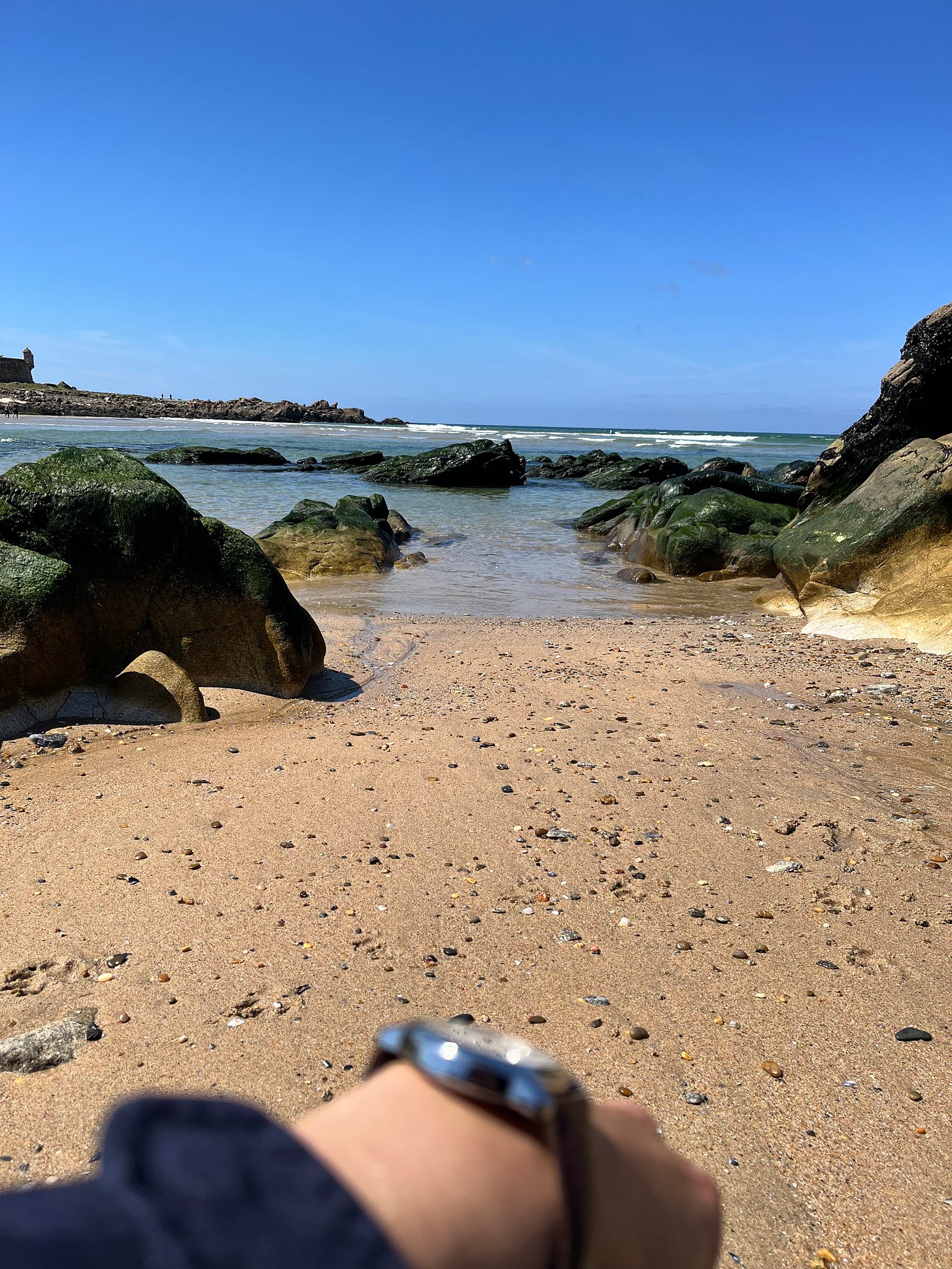 image: foreground with a hand wearing a watch, looking out to the beach and calm seas.