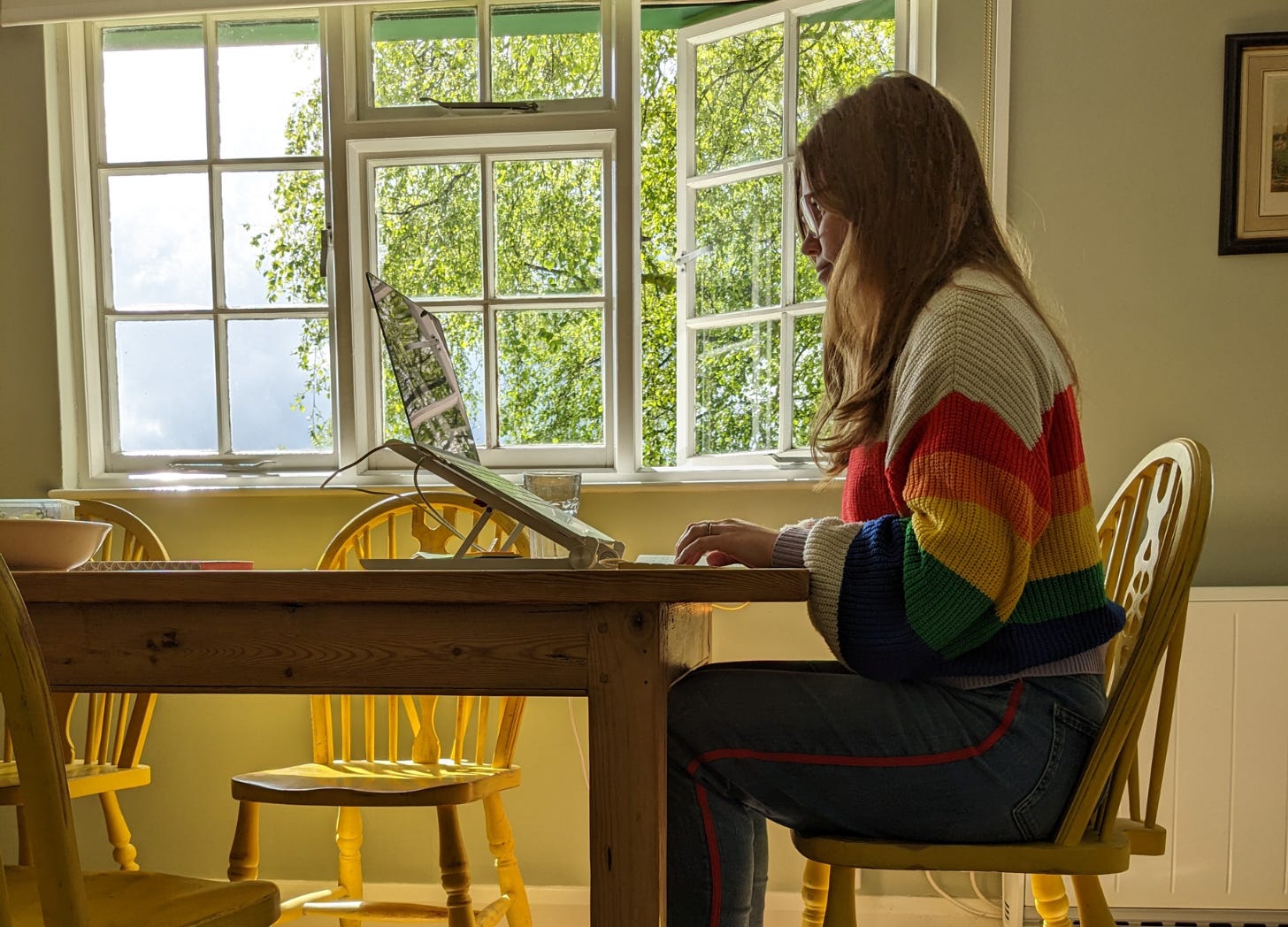 Libby Page is dressed in jeans and a rainbow jumper and sat at a table writing in a sunny room with a window and a view of a tree. 