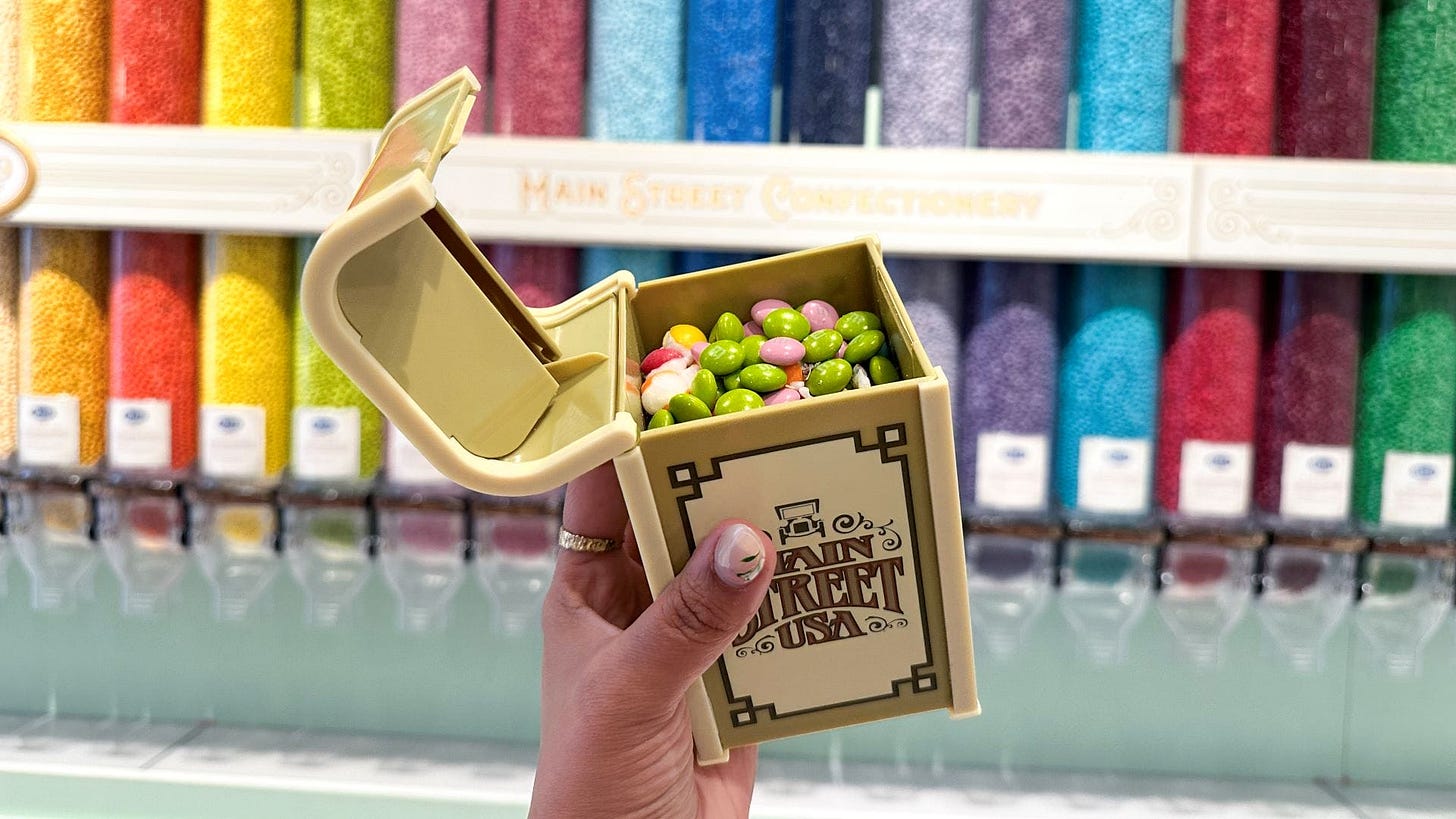 A hand holds an open candy box filled with colorful candies in front of candy dispensers on a display wall.