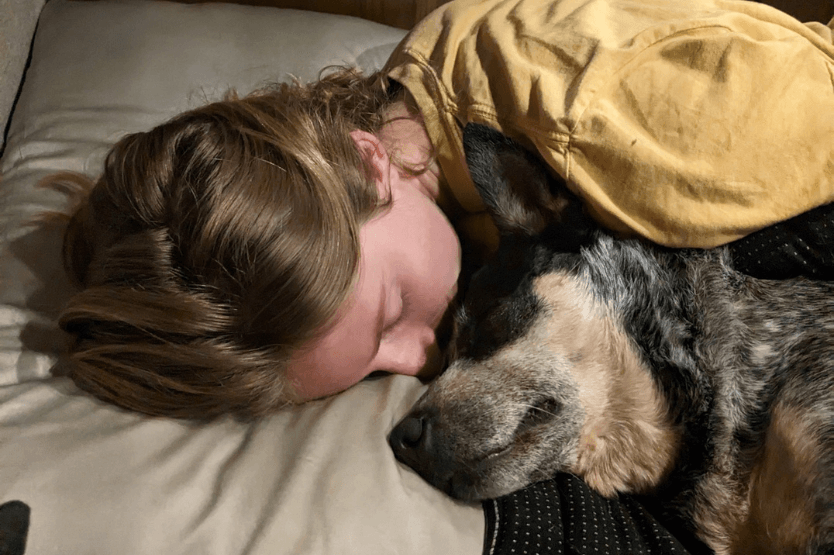 Haley, a young woman wearing a yellow shirt, snuggles closely against her Australian cattle dog, Scout, while in bed