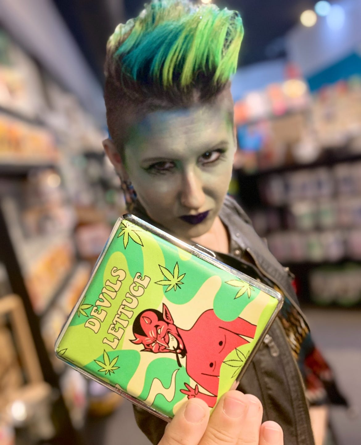Lyric, a few years ago at Halloween, dressed as a mer-creature, which later became the “weed creature” in their minds after this photo. They stand in a store holding up a wallet that says the Devil’a lettuce on it, with a red devil and green weed leaves. 