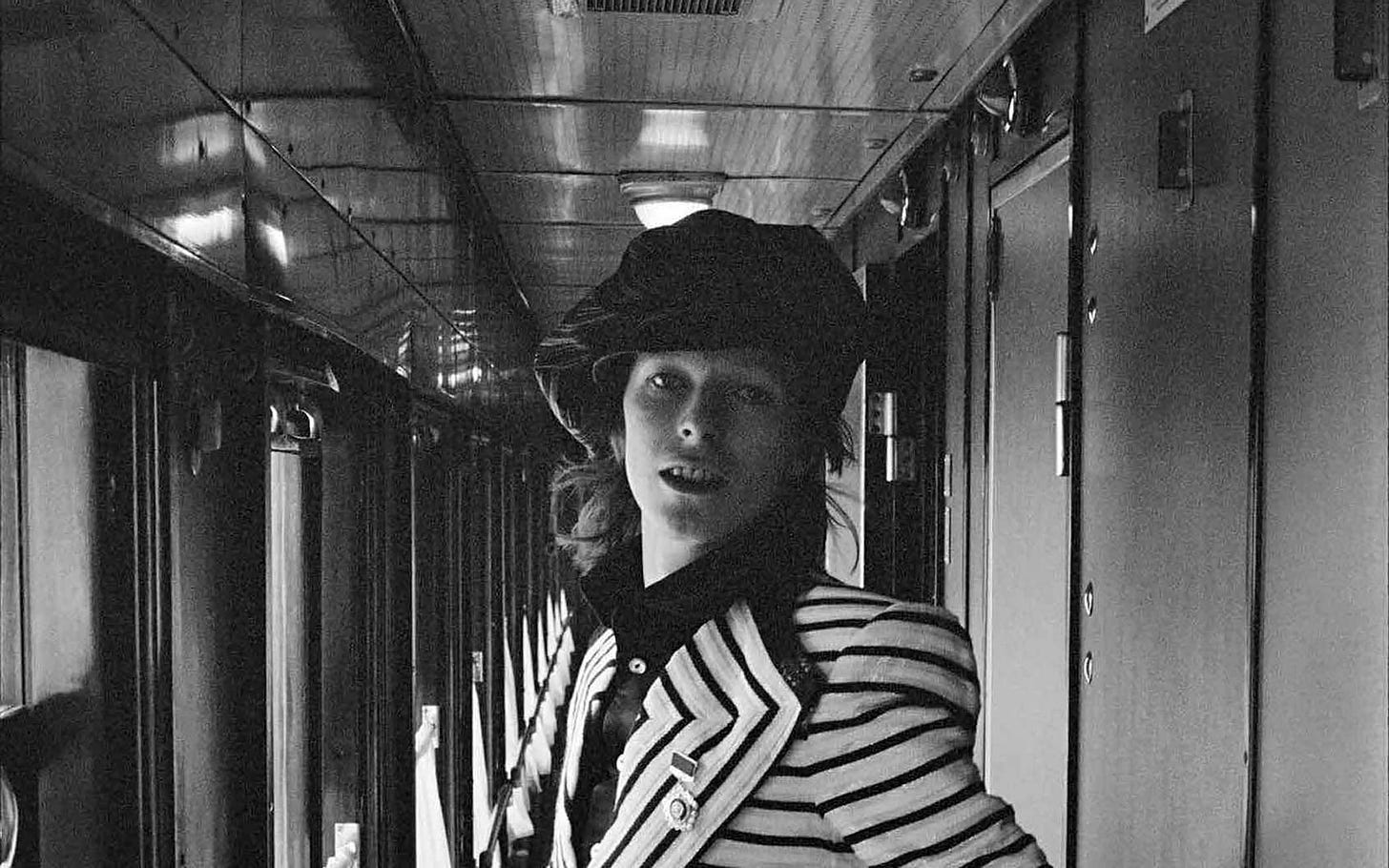 Foto preta e branca com David Bowie jovem no corredor de um trem. Ele usa boina e um terno listrado
