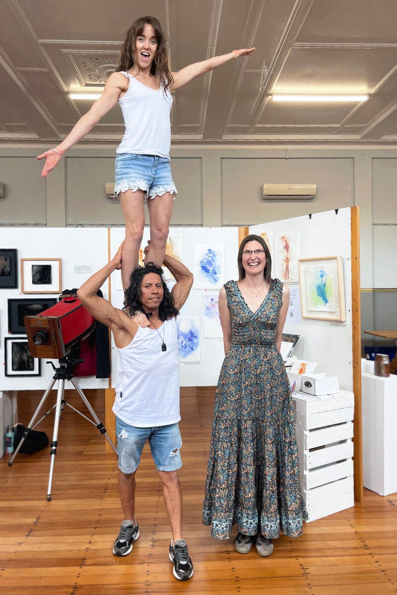 At The Hatch exhibition, Raglan Arts Weekend. On the left, a smiling woman stands on a man\'s shoulders with her arms outsretched "ta-da"! They are Jess and Jair (Jay) Ramirez, Raglan-based aerialist circus performers extraordinaire, co-founders of Freedom2Fly. On the right stands Ailene Cuthbertson, artist, smiling.