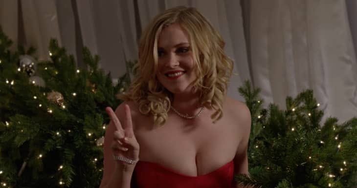 Movie still from Christmas Inheritance. A woman in a red party dress holds up a piece sign, Christmas lights and wreaths in the background.