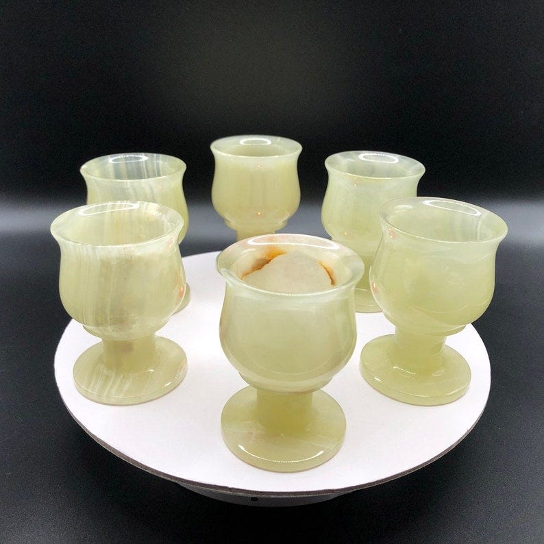 May include: Six green onyx shot glasses with a white interior. The glasses are arranged in a circle on a white surface.