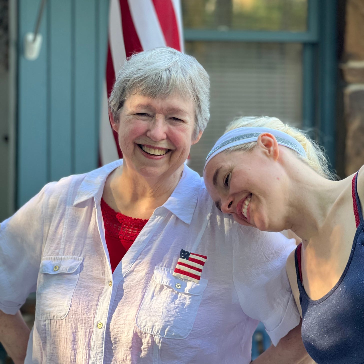 The author with her head on Mommy's shoulder, both grinning huge, identical grins, dressed in red-white-and-blue with the flag behind them, and another pinned to mom's blue shirt.